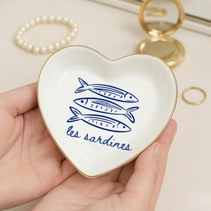 May include: A heart-shaped, white ceramic trinket dish with a gold rim. The dish features three blue fish illustrations and the text "les sardines" in blue script. The dish is held in a person's hands.