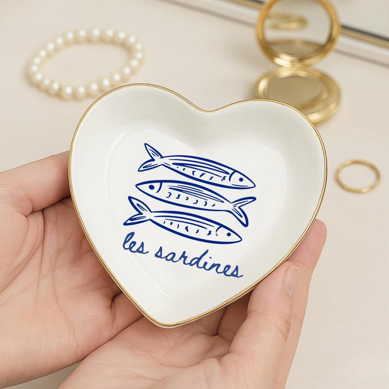 May include: A heart-shaped, white ceramic trinket dish with a gold rim. The dish features three blue fish illustrations and the text "les sardines" in blue script. The dish is held in a person's hands.
