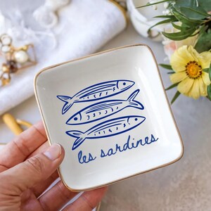May include: A square, white ceramic dish with a gold rim. The dish features three blue fish illustrations and the text "les sardines" in blue script. The dish is held by a hand.