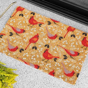 May include: A rectangular doormat with a repeating pattern of red cardinal birds on a yellow background with white floral accents. The birds are perched on black leafy branches. The mat is made of a textured material.
