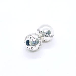 Może przedstawiać: Dwa srebrne charmsy Pandora. Każdy charms jest kulisty z opaską z przezroczystych kryształów. Jeden charms pokazuje wygrawerowany tekst "S925 ALE" i logo Pandora. Charmsy są na białym tle.