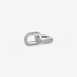 Puede incluir: Un charm plateado con dos eslabones rectangulares entrelazados. Uno de los eslabones está incrustado con pequeñas gemas transparentes en los bordes, mientras que el otro es liso. El charm está diseñado como un accesorio de joyería.