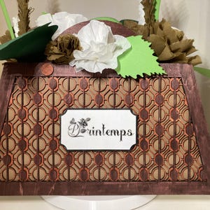 Può includere: Una scatola decorativa in legno con un motivo geometrico e la parola "Printemps" su un'etichetta bianca. La scatola è riempita di fiori di carta e foglie verdi. La scatola è marrone con accenti arancioni.