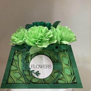Peut inclure: Une boîte à fleurs en bois vert avec un motif de feuilles superposées et le mot "FLOWERS" sur un cercle blanc. La boîte contient des fleurs en papier vert clair. L'ensemble est un arrangement floral décoratif.