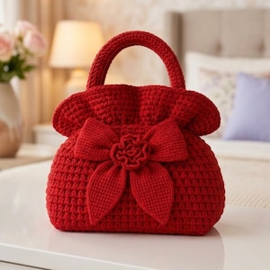 May include: A handmade red crochet handbag with a rounded shape and a top handle. The bag features a large bow with a flower detail on the front. The crochet stitches are visible, giving the bag a textured appearance.