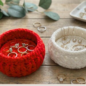 Peut inclure: Deux bols ronds crochet&eacute;s &agrave; la main pour le rangement de bijoux, l'un rouge vif et l'autre blanc cass&eacute;. Le bol rouge contient plusieurs bagues en or et argent avec diverses pierres. Le bol blanc contient des colliers, boucles et bracelets.