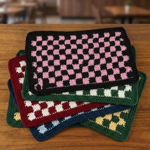May include: A stack of rectangular crochet mats featuring a chequered pattern. The top mat is black and pink, followed by layers in dark green and white, deep red and white, navy blue and light blue, and dark green and yellow colour combinations.