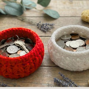 Peut inclure: Deux bols ronds crochet&eacute;s &agrave; la main avec un motif textur&eacute; en forme d'&eacute;toile. Le bol de gauche est rouge vif et contient un ensemble de cl&eacute;s en m&eacute;tal et de pi&egrave;ces de monnaie. Le bol de droite est blanc et rempli de pi&egrave;ces en argent et en cuivre.