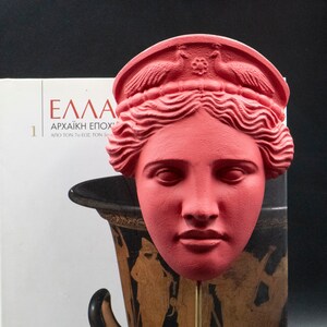 May include: A red plaster cast of a woman's face with a crown on her head. The face is on a black stand with a gold rod. The stand is in front of a book with the title "&Epsilon;&lambda;&lambda;ά&sigmaf; &Alpha;&rho;&chi;&alpha;ϊ&kappa;ή &Epsilon;&pi;&omicron;&chi;ή" on the cover.