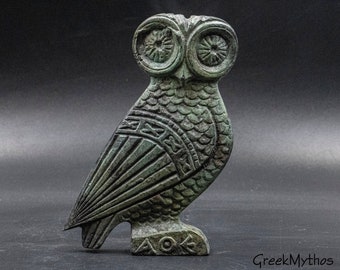 May include: A bronze-colored owl statue with detailed carvings. The owl has large, round eyes and intricate feather patterns. The base features Greek letters. The statue is displayed against a dark background.