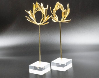May include: Two gold leaf sculptures of olive branches on clear acrylic stands. The sculptures are arranged symmetrically on a black background.