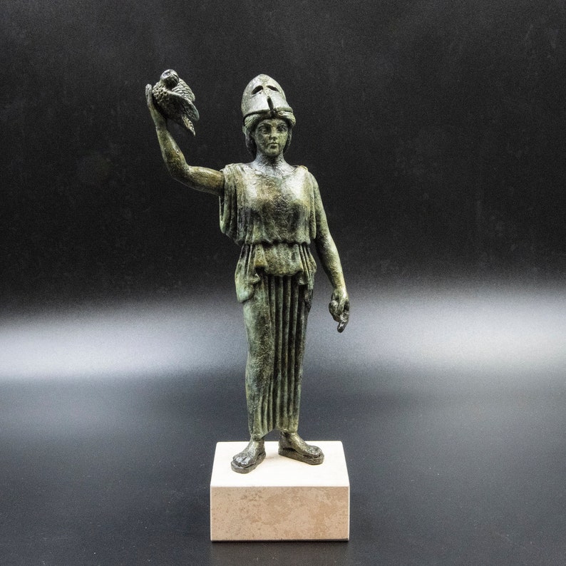 May include: A bronze statue of a woman wearing a helmet and holding a bird in her raised right hand. The statue is standing on a white pedestal.