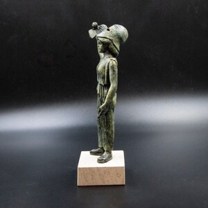 May include: A bronze statue of a woman wearing a helmet and holding a small object in her hand. The statue is standing on a square base.