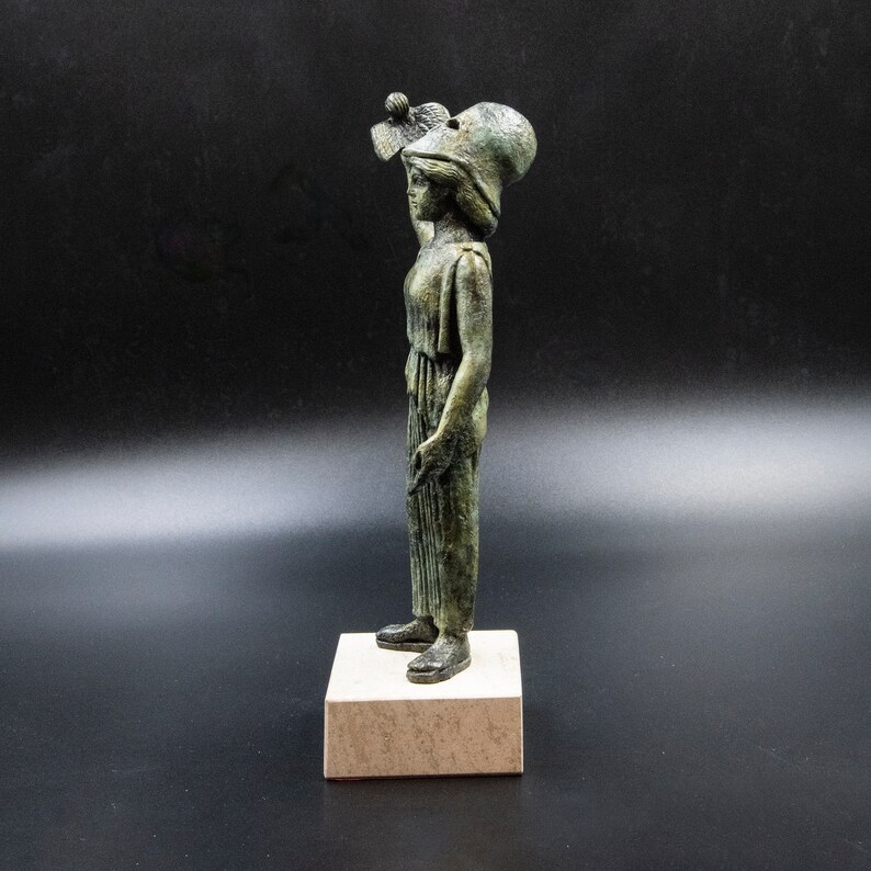 May include: A bronze statue of a woman wearing a helmet and holding a small object in her hand. The statue is standing on a square base.