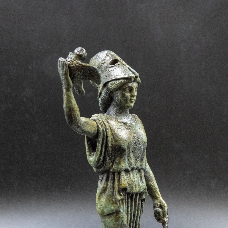 May include: A bronze statue of a female figure wearing a helmet and holding a bird in her right hand. The statue is standing on a black background.