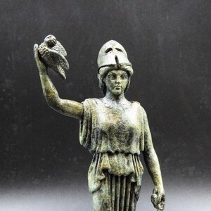 May include: A bronze statue of a woman wearing a helmet and holding a bird in her raised right hand. The statue is weathered and has a green patina.