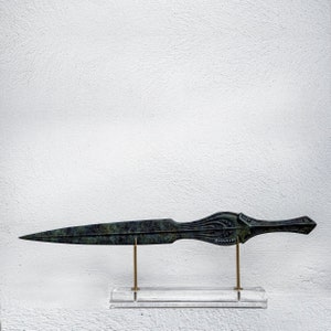 May include: A bronze dagger with a long, pointed blade and ornate handle. The handle features detailed carvings and a small inscription. The dagger is displayed on a clear acrylic stand with brass supports, against a white background.