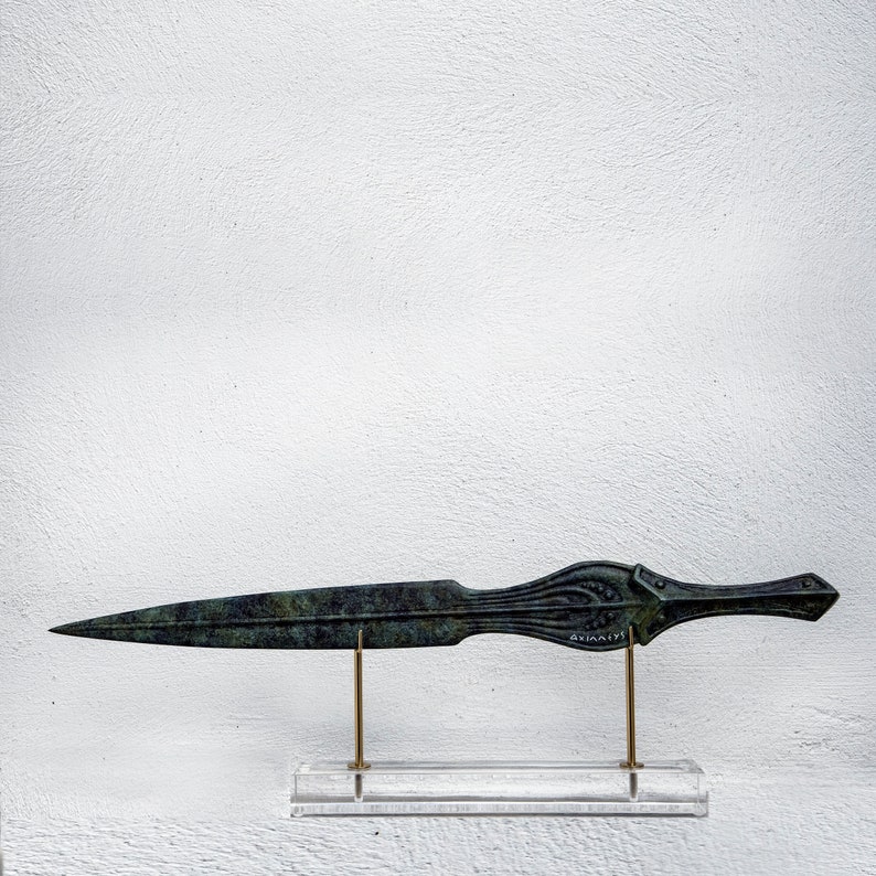 May include: A bronze dagger with a long, pointed blade and ornate handle. The handle features detailed carvings and a small inscription. The dagger is displayed on a clear acrylic stand with brass supports, against a white background.
