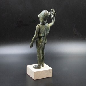 May include: A bronze statue of a figure wearing a helmet and holding a bird in its right hand. The figure is standing on a square base.