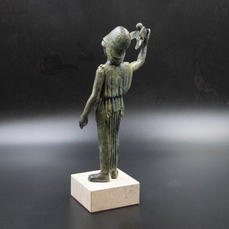 May include: A bronze statue of a figure wearing a helmet and holding a bird in its right hand. The figure is standing on a square base.