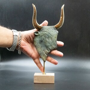 May include: A bronze bull's head sculpture with gold horns on a wooden base. The bull's head is detailed with intricate patterns and a textured surface.
