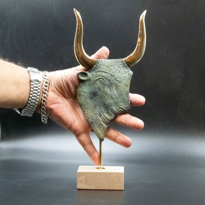 May include: A bronze bull's head sculpture with gold horns on a wooden base. The bull's head is detailed with intricate patterns and a textured surface.
