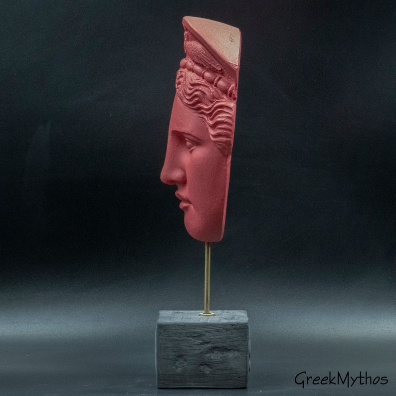 May include: A red painted plaster bust of a woman's face on a black wooden base with a gold metal stand. The bust is broken and only the face and top of the head are present. The woman is wearing a crown.