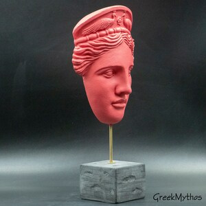 May include: A red plaster bust of a woman's head with a crown on top. The bust is on a black wooden base with a gold metal rod.