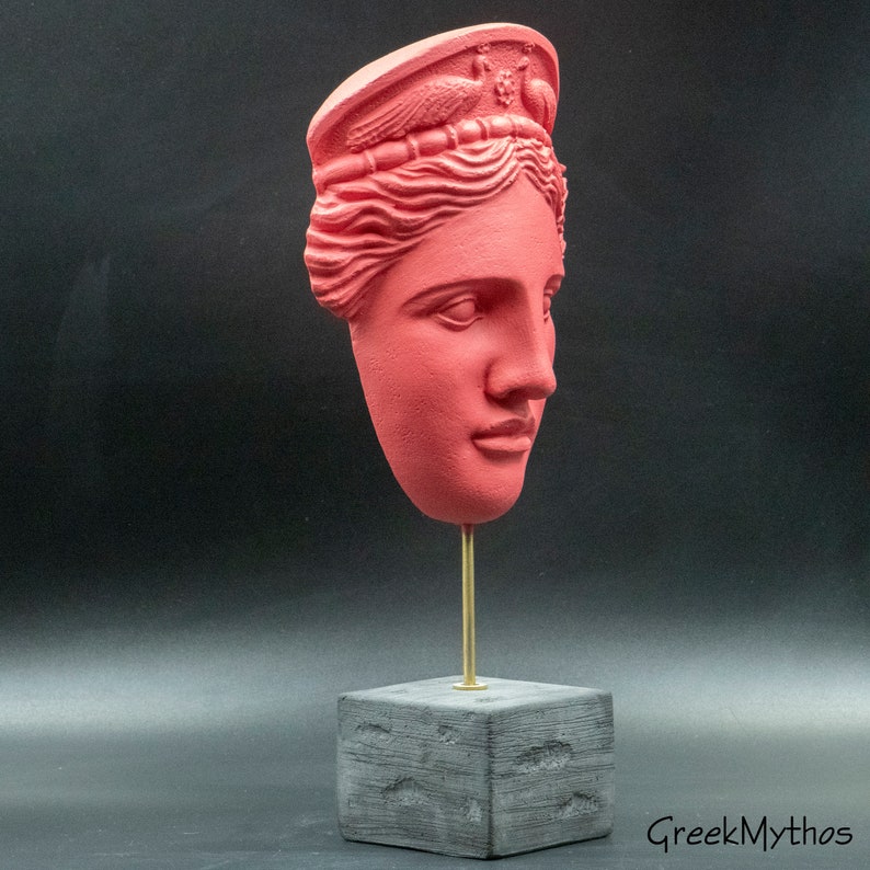 May include: A red plaster bust of a woman's head with a crown on top. The bust is on a black wooden base with a gold metal rod.