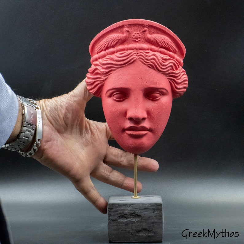 May include: A red painted plaster bust of a woman's head with a crown of peacocks on top. The bust is on a black wooden stand.