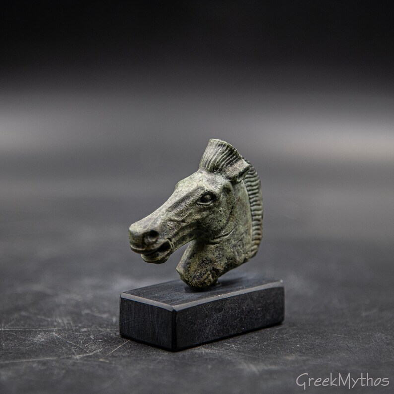 May include: A small, green bronze sculpture of a horse's head. The sculpture is on a black base.