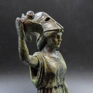 May include: A bronze statue of a woman wearing a helmet and holding a bird in her hand. The statue is weathered and has a green patina.