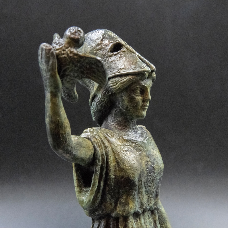 May include: A bronze statue of a woman wearing a helmet and holding a bird in her hand. The statue is weathered and has a green patina.