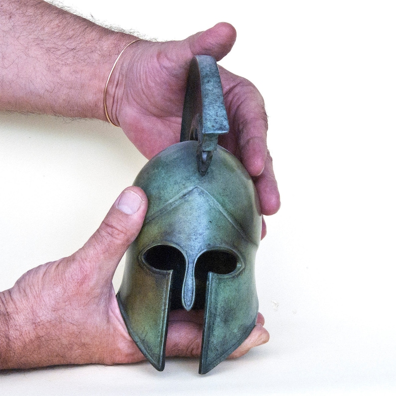May include: A bronze Greek warrior helmet with a plume on top. The helmet has a green patina and is decorated with a geometric design.