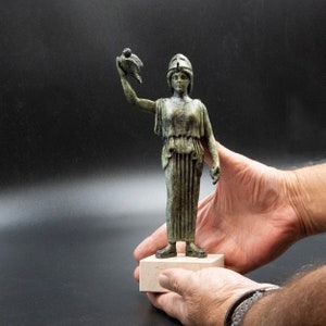 May include: A bronze statue of a woman holding a bird in her left hand. She is wearing a helmet and a long, flowing robe. The statue is on a white base.