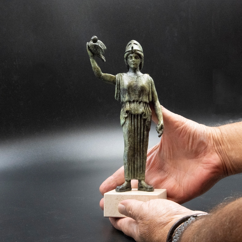 May include: A bronze statue of a woman holding a bird in her left hand. She is wearing a helmet and a long, flowing robe. The statue is on a white base.