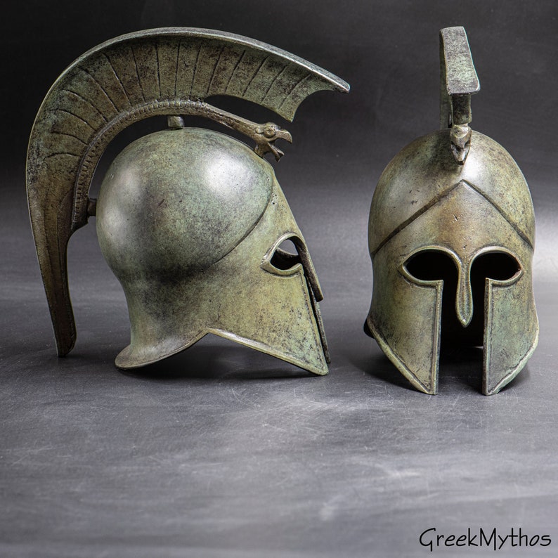 May include: Two antique bronze Greek warrior helmets with a plume on top. The helmet on the left has a winged crest. The helmet on the right has a simple crest.