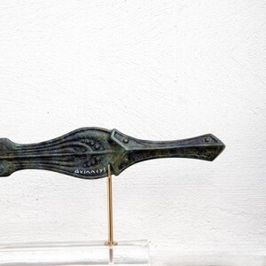 May include: Bronze-colored, decorative dagger with intricate floral and geometric designs. The dagger is mounted on a gold-colored stand and a clear base. The text "AXIANEVS" is inscribed on the dagger. The dagger is displayed against a white background.