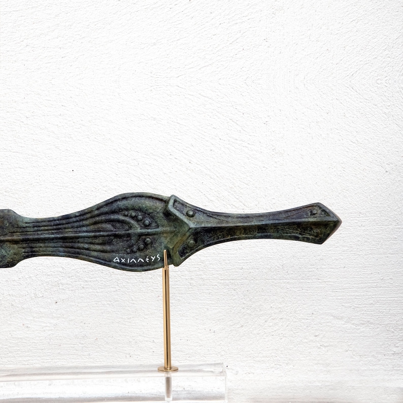May include: Bronze-colored, decorative dagger with intricate floral and geometric designs. The dagger is mounted on a gold-colored stand and a clear base. The text "AXIANEVS" is inscribed on the dagger. The dagger is displayed against a white background.