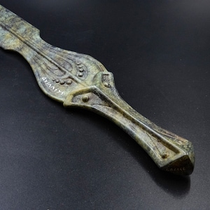May include: A detailed close-up of a green and bronze-colored decorative dagger. The blade features a stylized design with the Greek word "&Alpha;&Chi;&Iota;&Lambda;&Lambda;&Epsilon;&Upsilon;&Sigma;" inscribed. The handle is adorned with geometric patterns and the word "GREECE".