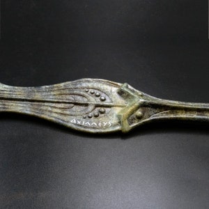 May include: Close-up of an antique bronze decorative object with an elongated, leaf-like shape. The object features intricate patterns of lines and small circular details, with the word "AXIANEVS" inscribed in white. The bronze has a weathered, aged appearance.