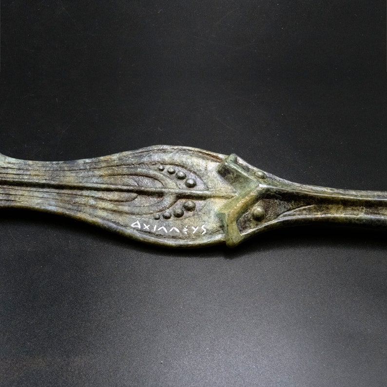 May include: Close-up of an antique bronze decorative object with an elongated, leaf-like shape. The object features intricate patterns of lines and small circular details, with the word "AXIANEVS" inscribed in white. The bronze has a weathered, aged appearance.