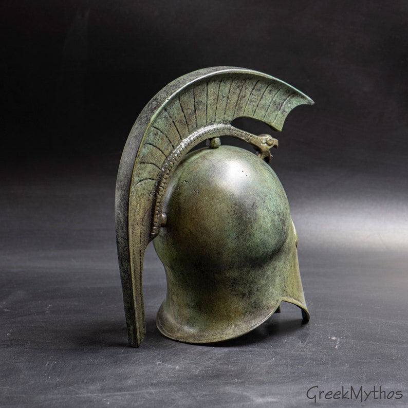 May include: A bronze Greek warrior helmet with a crested plume and a serpent design on the top. The helmet is weathered and has a green patina.