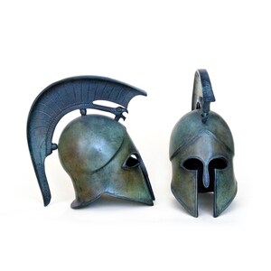 May include: Two bronze Greek warrior helmets, one with a plume and the other with a crest. The helmets are detailed with a face mask and a chin strap.