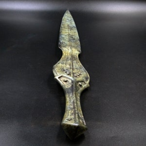 May include: A green and gold dagger with a pointed blade and ornate handle. The dagger has a textured surface and a geometric design on the handle. The blade is sharp and the overall design is detailed.