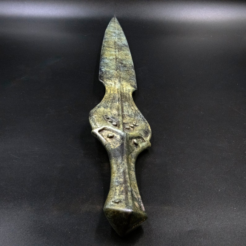 May include: A green and gold dagger with a pointed blade and ornate handle. The dagger has a textured surface and a geometric design on the handle. The blade is sharp and the overall design is detailed.