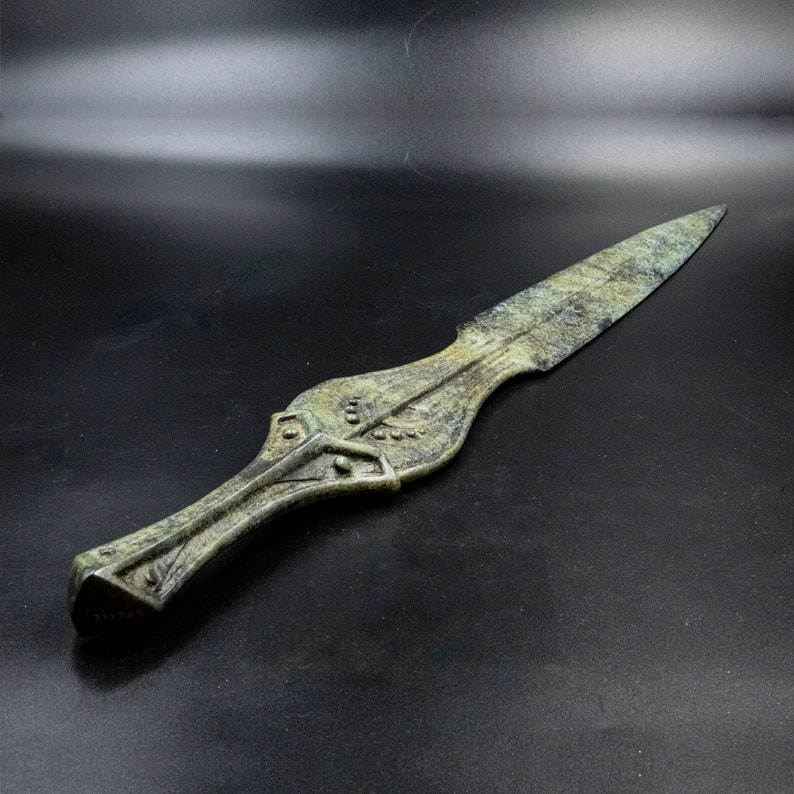 May include: An antique-style spearhead with a weathered, greenish-bronze finish. The spearhead features a long, pointed blade and a detailed handle with geometric and leaf-like patterns. The overall design suggests a historical or archaeological artifact.