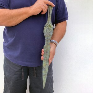 May include: A bronze-colored, elongated dagger with intricate decorative details. The dagger has a pointed blade and a handle with ornate carvings. The text "ARIANITI" is visible on the blade. The dagger is held against a white background.