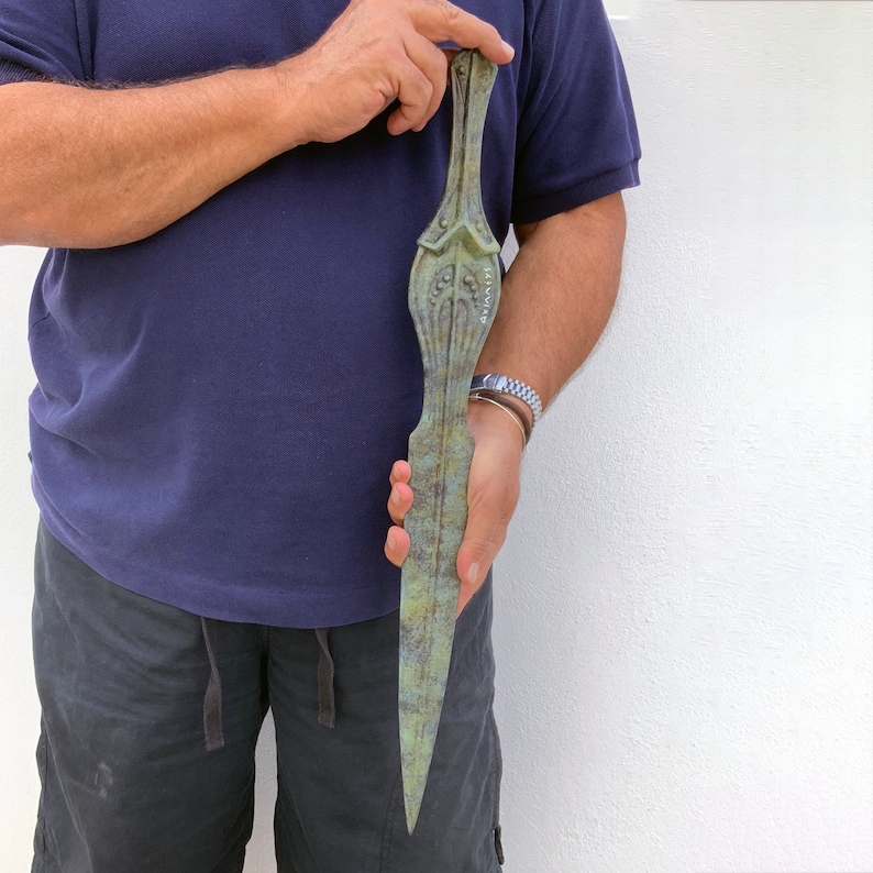 May include: A bronze-colored, elongated dagger with intricate decorative details. The dagger has a pointed blade and a handle with ornate carvings. The text "ARIANITI" is visible on the blade. The dagger is held against a white background.