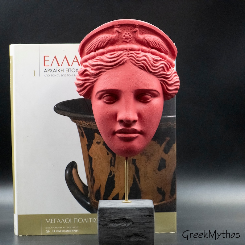 May include: A red plaster cast of a woman's face with a crown on her head. The face is on a black stand with a gold rod. The stand is in front of a book with the title "&Epsilon;&lambda;&lambda;ά&sigmaf; &Alpha;&rho;&chi;&alpha;ϊ&kappa;ή &Epsilon;&pi;&omicron;&chi;ή" on the cover.